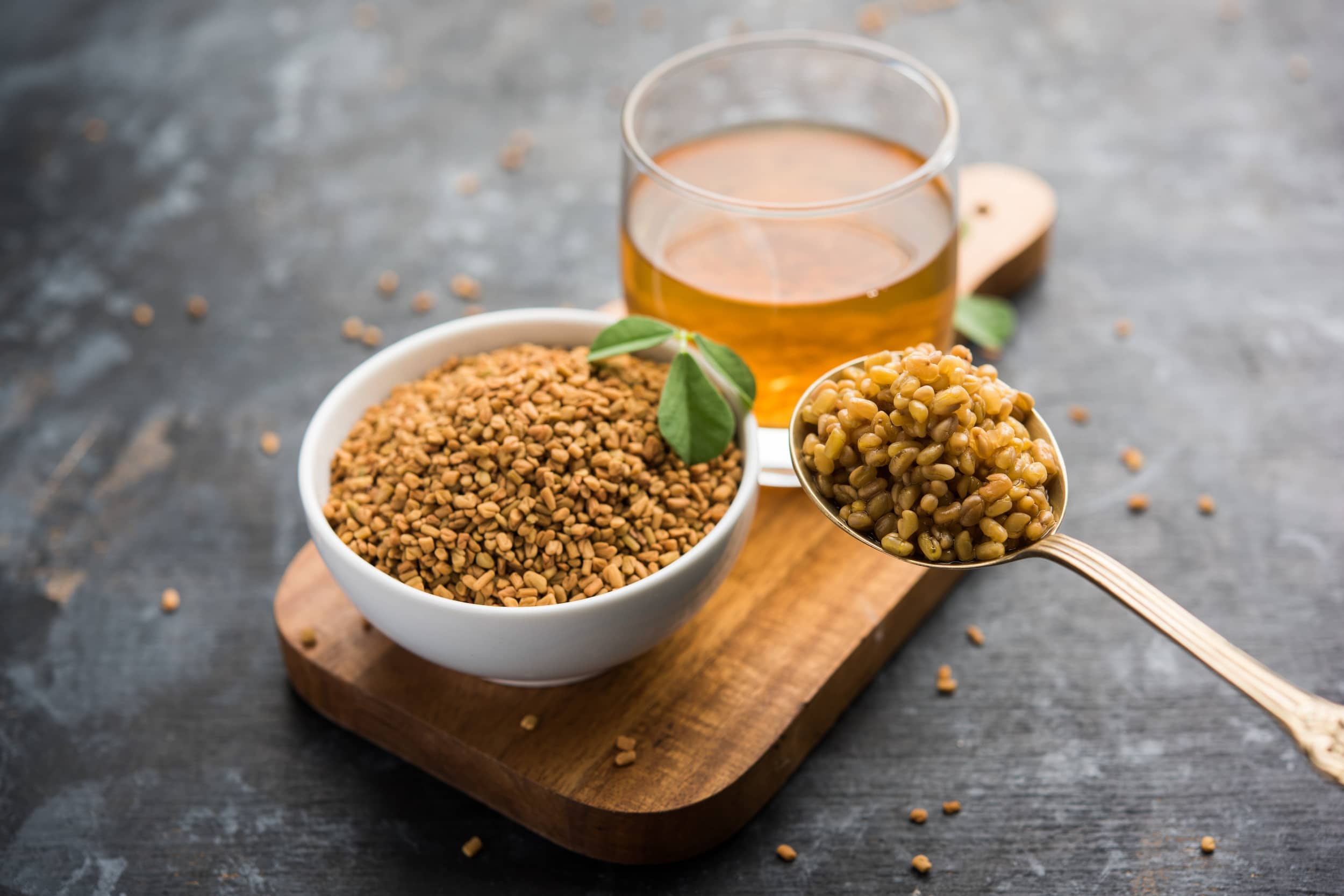 A spoon of fenugreek with a bowl and teacup of fenugreek in the background.